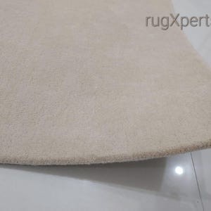 May include: Close-up of a beige rug with a soft, textured surface. The rug has a rounded edge and is likely made of a plush material. The rug is a neutral color, suitable for various interior design styles.