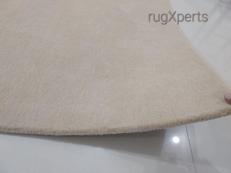 May include: Close-up of a beige rug with a soft, textured surface. The rug has a rounded edge and is likely made of a plush material. The rug is a neutral color, suitable for various interior design styles.