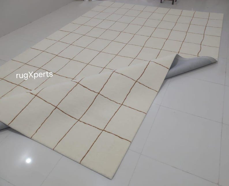 May include: A white rug with a grid pattern of brown lines. The rug is rolled up and the backing is visible.