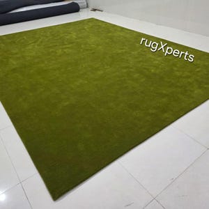 May include: A large, rectangular, olive green rug. The rug has a soft, plush texture and is displayed on a tiled floor. The text "rugXperts" is visible in the upper right corner.