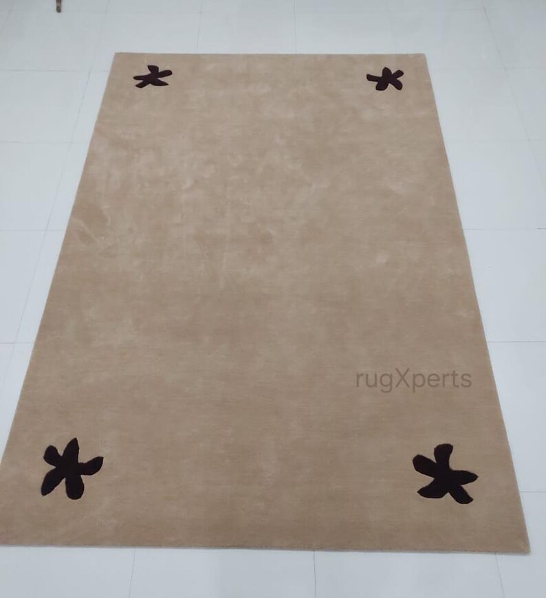 May include: A rectangular beige rug with four dark brown star-shaped designs evenly spaced on the corners. The rug has a soft, plush texture and is suitable for living rooms or bedrooms.