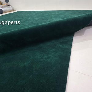 May include: A large, emerald green rug with a soft, velvety texture. The rug is partially rolled up, revealing a dark grey backing. The text "rugXperts" is visible in the upper left corner. The rug is on a white tiled floor.