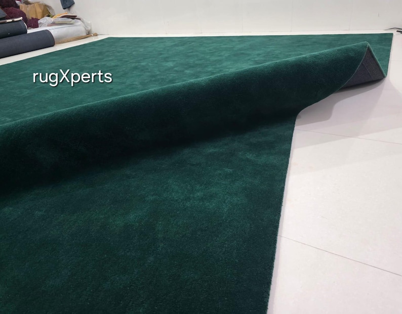 May include: A large, emerald green rug with a soft, velvety texture. The rug is partially rolled up, revealing a dark grey backing. The text "rugXperts" is visible in the upper left corner. The rug is on a white tiled floor.