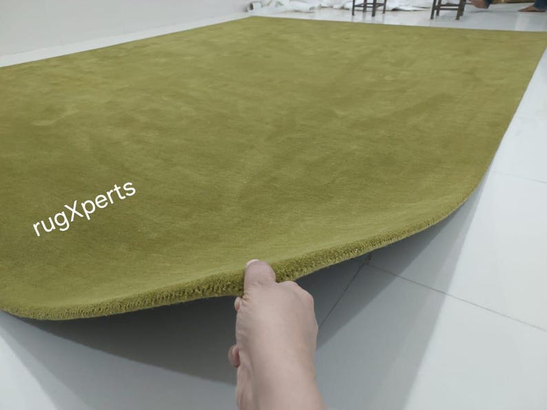 May include: A large, rectangular olive green rug. The rug's edge is being lifted, revealing its thickness and texture. The text "rugXperts" is visible in the corner. This rug is suitable for living rooms and bedrooms.