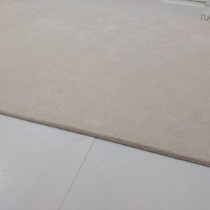 May include: A large, rectangular beige rug with a soft, textured surface. The rug is placed on a white tiled floor. The edge of the rug is a darker shade of beige. The rug is a neutral color and would complement many interior design styles.
