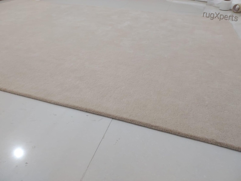 May include: A large, rectangular beige rug with a soft, textured surface. The rug is placed on a white tiled floor. The edge of the rug is a darker shade of beige. The rug is a neutral color and would complement many interior design styles.