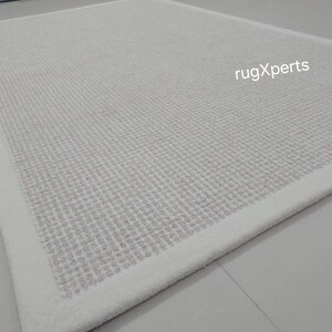 May include: A white rug with a subtle textured pattern and a white border. The rug is made of a soft, comfortable material and is perfect for adding a touch of elegance to any room.