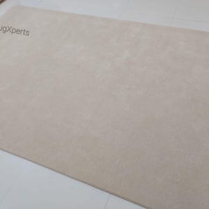 May include: A large, rectangular beige rug. The rug has a soft, plush texture and is a solid color. The words "rugXperts" are printed in the upper left corner. The rug is on a white tiled floor.
