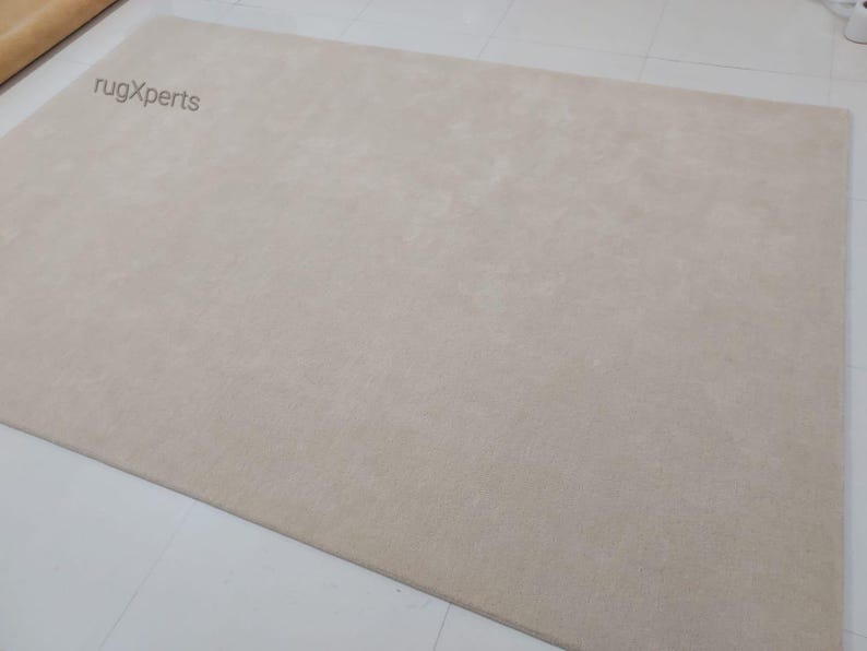 May include: A large, rectangular beige rug. The rug has a soft, plush texture and is a solid color. The words "rugXperts" are printed in the upper left corner. The rug is on a white tiled floor.