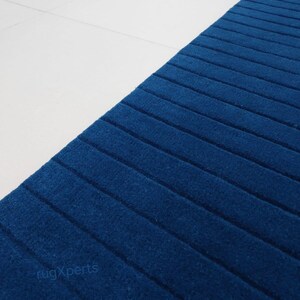 Op de afbeelding: Een close-up van een blauw tapijt met een geribbelde textuur, afgewisseld met witte tegels. Het tapijt is diepblauw, met parallelle lijnen die een gestructureerd patroon vormen. De tekst "rugXperts" is zichtbaar.