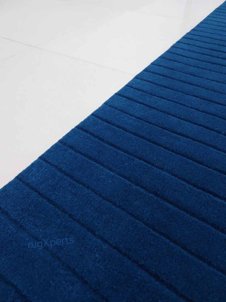 Op de afbeelding: Een close-up van een blauw tapijt met een geribbelde textuur, afgewisseld met witte tegels. Het tapijt is diepblauw, met parallelle lijnen die een gestructureerd patroon vormen. De tekst "rugXperts" is zichtbaar.