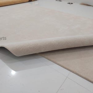 May include: Beige rug with a rolled edge, revealing the underside. The rug is on a white tiled floor. The text "rugXperts" is visible in the corner. The rug appears to be made of a soft, plush material.
