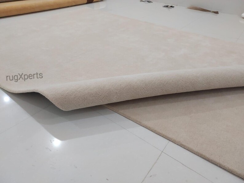 May include: Beige rug with a rolled edge, revealing the underside. The rug is on a white tiled floor. The text "rugXperts" is visible in the corner. The rug appears to be made of a soft, plush material.