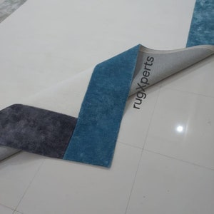 May include: A rectangular rug with a geometric design. The rug features a white central field with sections of blue, grey, and brown. The rug's edge is folded back, revealing the underside and the text "rugXperts".