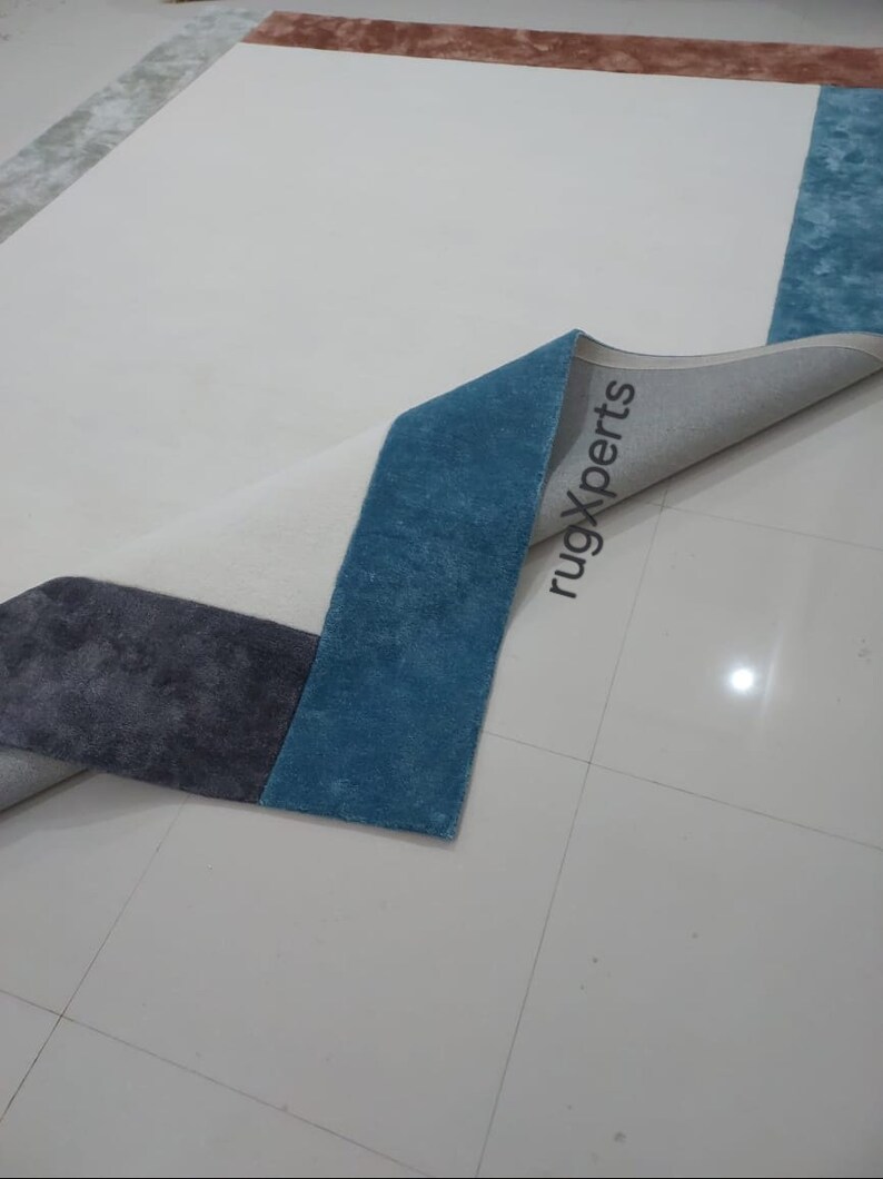 May include: A rectangular rug with a geometric design. The rug features a white central field with sections of blue, grey, and brown. The rug's edge is folded back, revealing the underside and the text "rugXperts".
