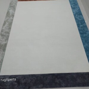 May include: A large rectangular rug with a white central area and borders in grey, brown, and blue. The rug has a plush texture and a modern design, suitable for a living room or bedroom. The rug's dimensions are not visible.