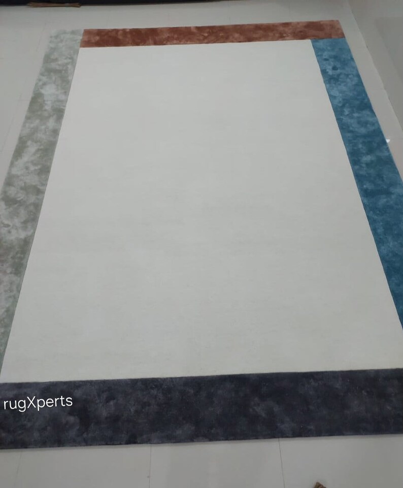 May include: A large rectangular rug with a white central area and borders in grey, brown, and blue. The rug has a plush texture and a modern design, suitable for a living room or bedroom. The rug's dimensions are not visible.