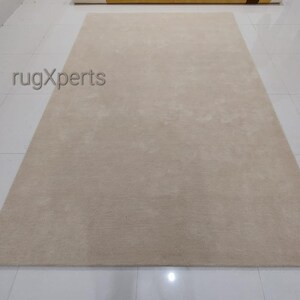 May include: A large, rectangular beige rug with a soft, textured surface. The rug is placed on a white tiled floor. The text "rugXperts" is visible in the upper left corner.