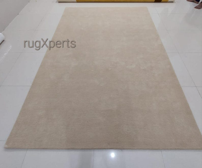 May include: A large, rectangular beige rug with a soft, textured surface. The rug is placed on a white tiled floor. The text "rugXperts" is visible in the upper left corner.