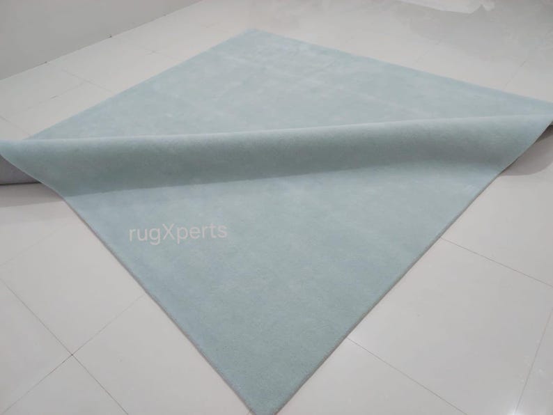 May include: A light blue rectangular rug with a rolled edge. The rug is made of a soft material and is displayed on a white tiled floor. The text "rugXperts" is visible on the rug.