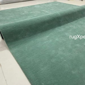May include: A rolled-up, teal-coloured rug. The rug is made of a soft material and is partially unrolled, revealing its texture. The text "rugXperts" is visible on the rug's surface.