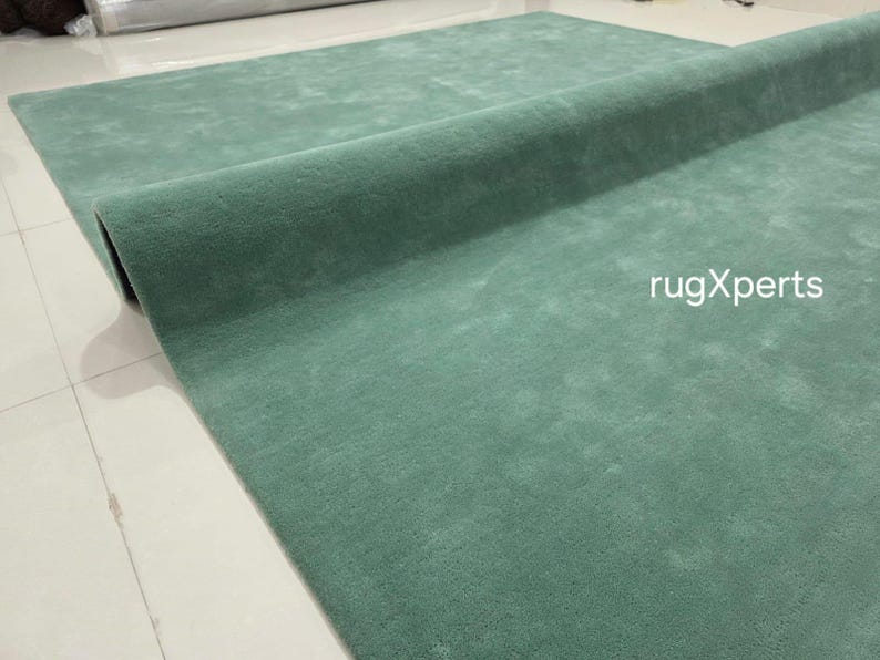 May include: A rolled-up, teal-coloured rug. The rug is made of a soft material and is partially unrolled, revealing its texture. The text "rugXperts" is visible on the rug's surface.