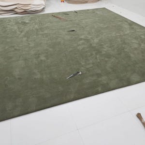 May include: A large, olive green rug with a soft, textured appearance. The rug is rectangular and covers a significant portion of the floor. Several small objects are scattered across the surface, and a broom is visible in the corner. The rug is a home decor item.