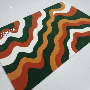 May include: A rectangular rug with a wavy pattern in shades of green, white, orange, and rust. The rug features the text "rugXperts" in the upper left corner. The design is a modern, abstract style.