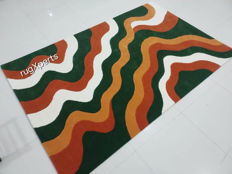 May include: A rectangular rug with a wavy pattern in shades of green, white, orange, and rust. The rug features the text "rugXperts" in the upper left corner. The design is a modern, abstract style.