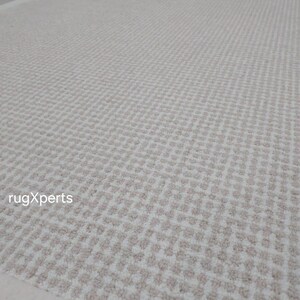 May include: A close-up of a white and beige patterned rug with a subtle geometric design. The rug has a white border and is made of a soft, textured material.