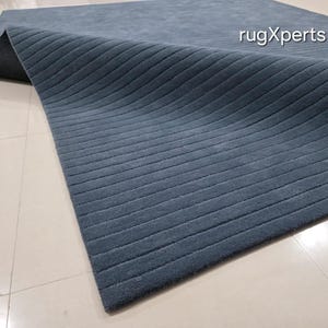 Op de afbeelding: Een blauw tapijt met een geribbelde textuur. Het tapijt is gedeeltelijk opgerold en onthult een donkere onderkant. Het tapijt ligt op een lichtgekleurde tegelvloer. De tekst "rugXperts" is zichtbaar op het tapijt.