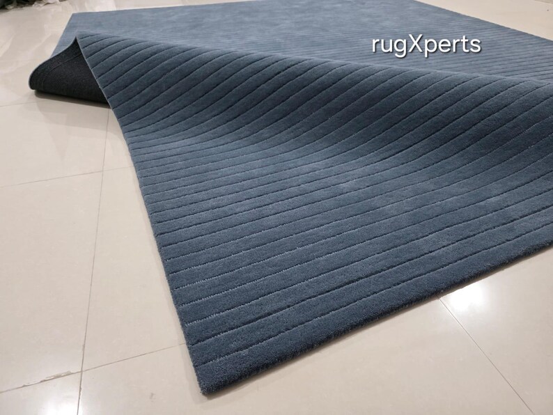 Op de afbeelding: Een blauw tapijt met een geribbelde textuur. Het tapijt is gedeeltelijk opgerold en onthult een donkere onderkant. Het tapijt ligt op een lichtgekleurde tegelvloer. De tekst "rugXperts" is zichtbaar op het tapijt.