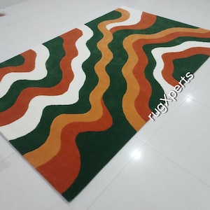 May include: A rectangular rug with a wavy pattern in shades of green, white, orange, and rust. The rug's design features flowing, organic lines that create a visually dynamic and modern aesthetic. The rug is placed on a white tiled floor.