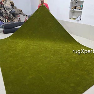 May include: A large, olive green rug is displayed, showcasing its plush texture and vibrant colour. The rug is being held up, revealing its size and shape. The text "rugXperts" is visible on the rug.