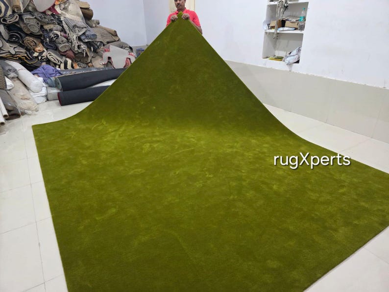 May include: A large, olive green rug is displayed, showcasing its plush texture and vibrant colour. The rug is being held up, revealing its size and shape. The text "rugXperts" is visible on the rug.