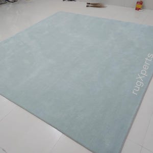 May include: A large, rectangular, light blue rug. The rug has a soft, plush texture and is placed on a white tiled floor. The text "rugXperts" is printed in white on the rug.