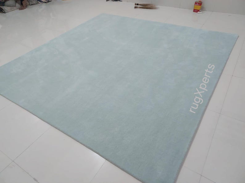 May include: A large, rectangular, light blue rug. The rug has a soft, plush texture and is placed on a white tiled floor. The text "rugXperts" is printed in white on the rug.