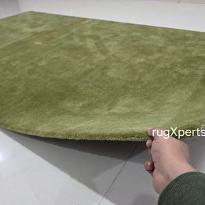 May include: A light green, semi-circular rug with a soft, textured surface. The rug is a solid colour and appears to be made of a plush material. The edge of the rug is slightly curved. The text "rugXperts" is visible.