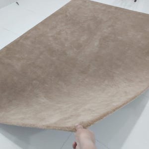 May include: A large, beige-colored rug with a soft, plush texture. The rug has a rounded shape and is displayed on a white tiled floor. The rug's fibers create a subtle, tonal variation in the beige color.