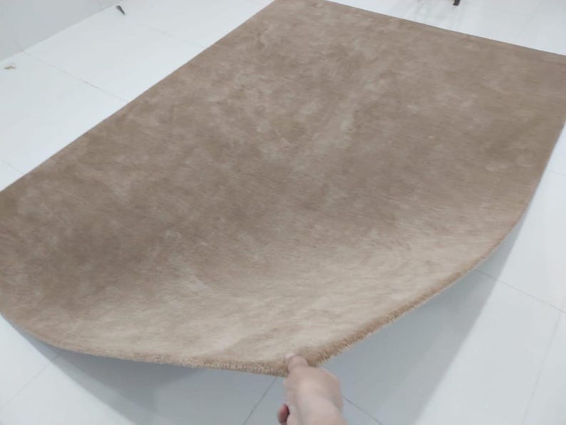 May include: A large, beige-colored rug with a soft, plush texture. The rug has a rounded shape and is displayed on a white tiled floor. The rug's fibers create a subtle, tonal variation in the beige color.