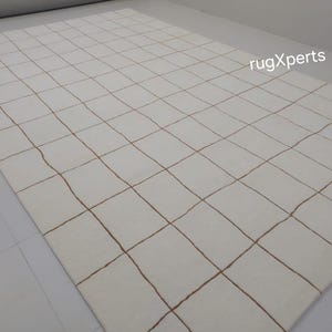 May include: White rug with a grid pattern of thin brown lines. The rug is made of a soft, woven material.