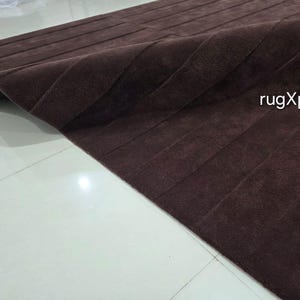 May include: A brown rectangular rug with a textured, striped design. The rug is partially folded, revealing a dark underside. The text "rugXperts" is visible in the image.