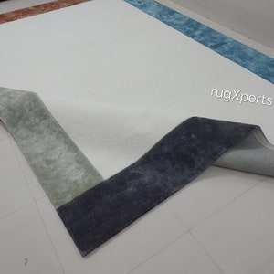 May include: A large, rectangular rug with a white central area and a multi-coloured border. The border features sections of blue, rust, and grey-green. The rug's corner is folded back, revealing the backing. The text "rugXperts" is visible.