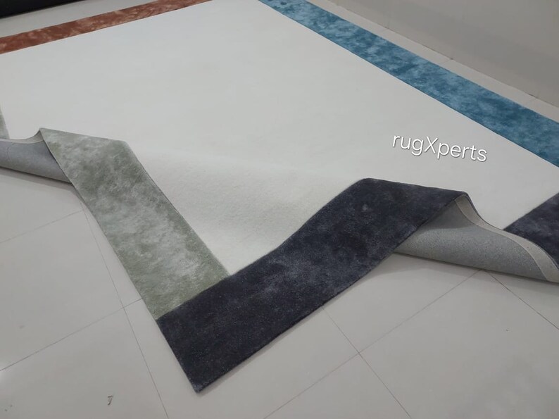 May include: A large, rectangular rug with a white central area and a multi-coloured border. The border features sections of blue, rust, and grey-green. The rug's corner is folded back, revealing the backing. The text "rugXperts" is visible.