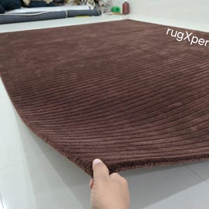 Op de afbeelding: Een groot, rechthoekig bruin tapijt met een geribbelde textuur. Het tapijt is gemaakt van een zacht materiaal en heeft een gebogen rand. De tekst "rugXperts" is zichtbaar in de rechterbovenhoek.