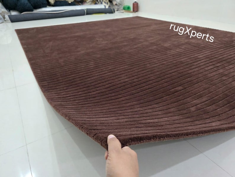 Op de afbeelding: Een groot, rechthoekig bruin tapijt met een geribbelde textuur. Het tapijt is gemaakt van een zacht materiaal en heeft een gebogen rand. De tekst "rugXperts" is zichtbaar in de rechterbovenhoek.