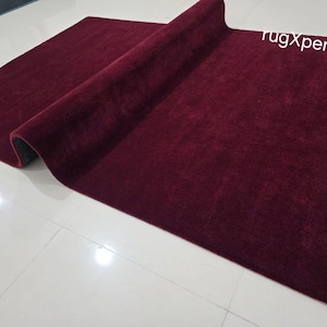 May include: A rolled-up, rectangular burgundy rug with a plush texture. The rug is partially unrolled, revealing a dark gray underside. The words "rugXperts" are visible in the upper right corner.