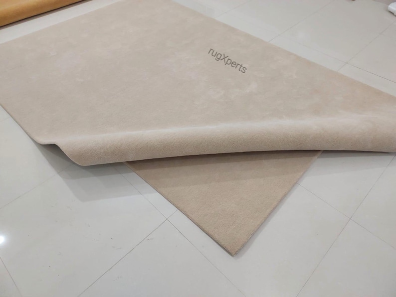 May include: Beige rug with a rolled edge, partially covering a second rug of the same color. The text "rugXperts" is visible on the top rug. The rugs are on a tiled floor.