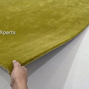 May include: A close-up of a section of a green rug. The rug has a soft, plush texture and a solid colour. The text "rugXperts" is in the upper left corner. The rug is on a light grey surface.