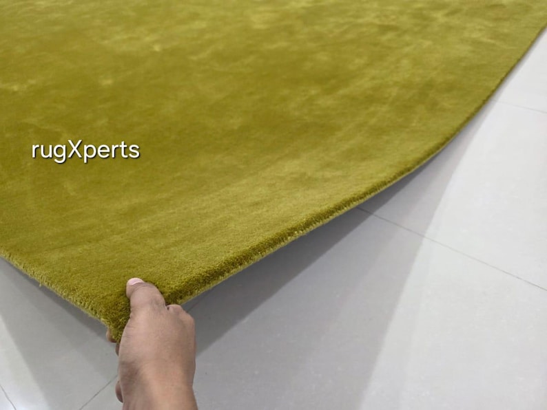 May include: A close-up of a section of a green rug. The rug has a soft, plush texture and a solid colour. The text "rugXperts" is in the upper left corner. The rug is on a light grey surface.